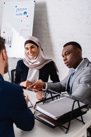 african american businessman pointing with finger near smiling arabian businesswoman and translator on blurred foregroundの写真素材