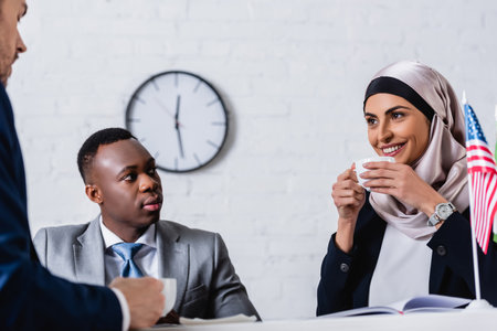 cheerful arabian businesswoman drinking coffee near multicultural business partners and american flag on blurred foregroundの写真素材