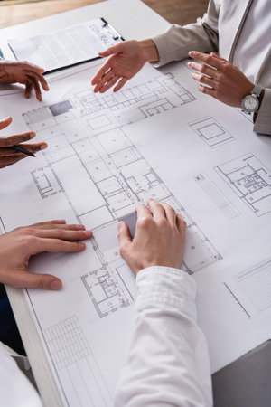 cropped view of businesswoman pointing with hand at blueprint near multiethnic business partners