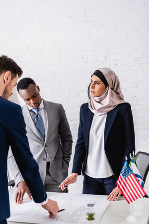 serious arabian businesswoman pointing with finger near blueprint during meeting with interracial business partnersの写真素材