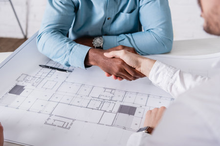 cropped view of african american businessman shaking hands with business partner near blueprint, blurred foregroundの写真素材