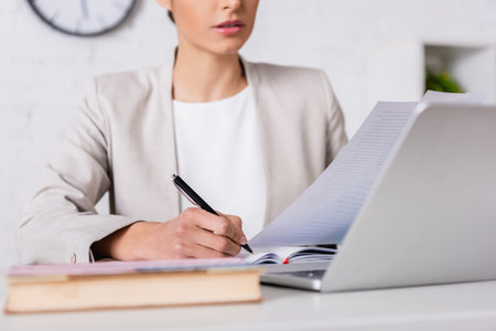 cropped view of translator writing in notebook while holding document near laptop, blurred foregroundの写真素材