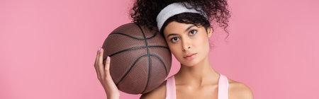 curly young woman holding basketball isolated on pink, horizontal bannerの写真素材