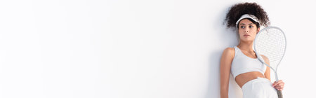 curly sportswoman in cap looking away while leaning on wall and holding tennis racket on white, bannerの写真素材