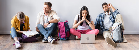 Multiethnic students with laptop, book and backpacks sitting on floor, bannerの写真素材