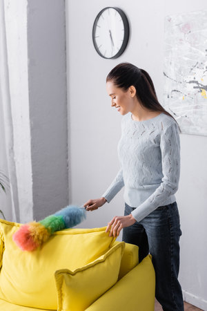 smiling woman cleaning yellow sofa with dust brush at homeの写真素材