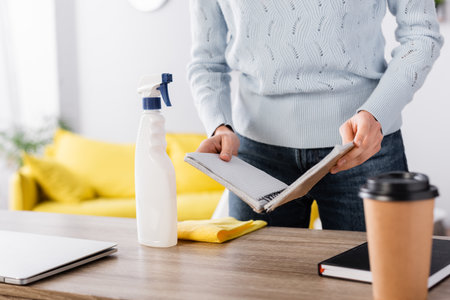partial view of housewife holding notebook near rag, detergent, and coffee to go on blurred...