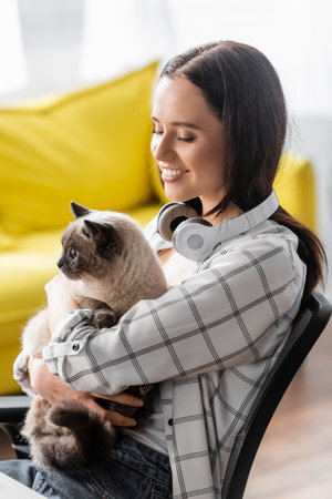 smiling woman with headphones on neck embracing cat at homeの写真素材