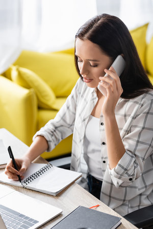 young freelancer writing in notebook and talking on smartphone at homeの写真素材