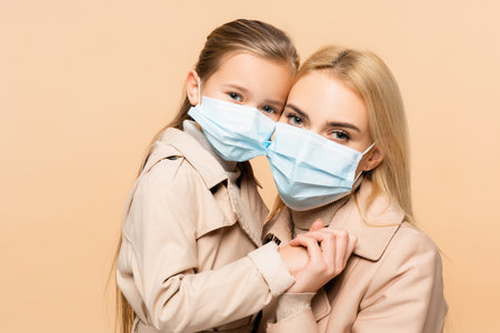 mother and daughter in medical masks holding hands isolated on beigeの写真素材