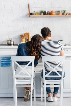 back view of woman leaning on man and sitting on chair in kitchenの写真素材