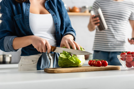 partial view of woman cutting lettuce near ingredients on kitchen table and boyfriend on blurred backgroundの写真素材