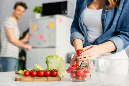 cropped view of woman holding red cherry tomato near ingredients on kitchen table and boyfriend on blurred backgroundの写真素材
