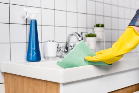 Cropped view of woman in rubber glove cleaning bathroom sink with rag near bottle of detergent on...