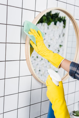 Cropped view of woman in yellow rubber gloves cleaning mirror with detergent and rag in bathroomの写真素材