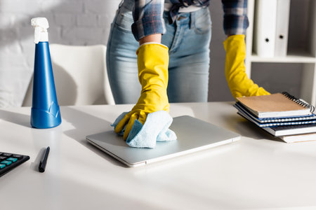 Cropped view of woman in rubber gloves cleaning laptop near detergent and notebooks on blurred background on tableの写真素材