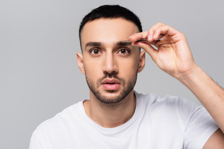 brunette hispanic man looking at camera while tweezing eyebrows isolated on grayの写真素材