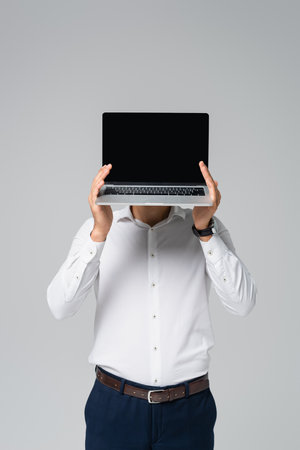 hispanic manager in white shirt obscuring face with laptop isolated on grayの写真素材