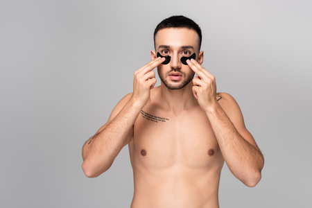young shirtless hispanic man applying eye patches isolated on grayの写真素材