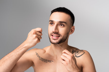 tattooed shirtless hispanic man looking away while holding dental floss isolated on grayの写真素材