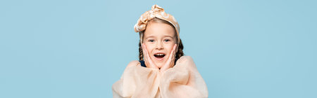 amazed kid in headband with bow touching cheeks isolated on blue, banner