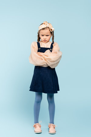 full length of offended kid with pigtails standing with crossed arms on blue