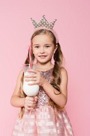 cheerful little girl in dress and crown holding glass with milkshake isolated on pinkの写真素材