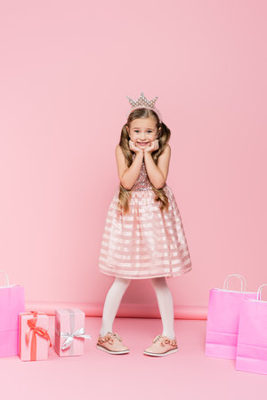 full length of excited little girl in crown standing near presents and shopping bags on pinkの写真素材