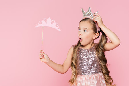 shocked little girl in dress holding paper crown on stick isolated on pinkの写真素材