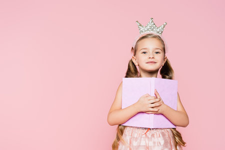 smiling little girl in crown holding book while looking at camera isolated on pinkの写真素材
