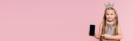 happy little girl in crown pointing with finger at smartphone with blank screen isolated on pink,...