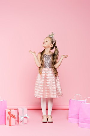 full length of pleased little girl in crown with closed eyes standing near presents and shopping bags on pinkの写真素材