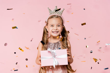 happy little girl in crown holding present near falling confetti on pinkの写真素材