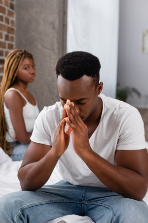 Depressed african american man sitting near girlfriend on blurred background in bedroomの写真素材