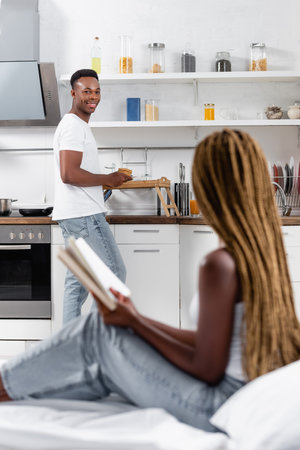 Smiling african american man holding pancakes near tray and girlfriend with book on blurred foreground on bedの写真素材