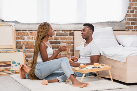 African american couple in jeans holding glasses of orange juice near pancakes on tray on floor in bedroomの写真素材