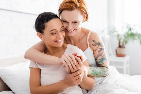 happy redhead lesbian woman with wedding ring on finger embracing african american girlfriend holding jewelry boxの写真素材