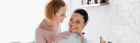 cheerful interracial lesbian couple smiling while looking at each other at home, bannerの写真素材