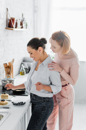 lesbian woman hugging shoulders of happy african american girlfriend serving breakfast in kitchenの写真素材