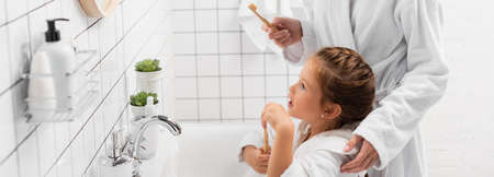 Kid in bathrobe holding toothbrush near mother and sink in bathroom, bannerの写真素材