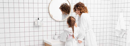Smiling woman in bathrobe looking at daughter near sink in bathroom, banner
