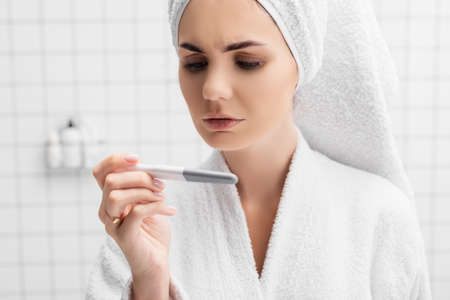 Worried woman in bathrobe looking at pregnancy test in modern bathroom