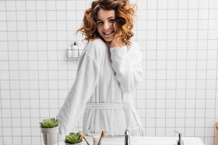 Smiling woman in white bathrobe standing near sink in bathroomの写真素材