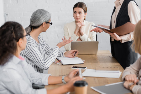 team leader looking at asian businesswoman near colleagues during meeting in officeの写真素材