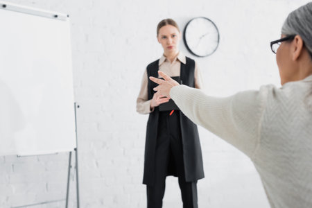 Asian businesswoman in glasses pointing with hand at flipchart near speaker with notebook on blurred backgroundの写真素材