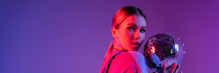 young and stylish woman holding shiny disco ball isolated on purple, banner