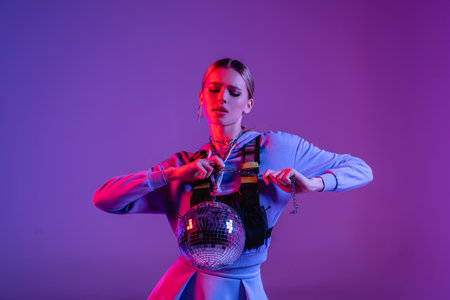 young and stylish woman posing with shiny disco ball on purpleの写真素材