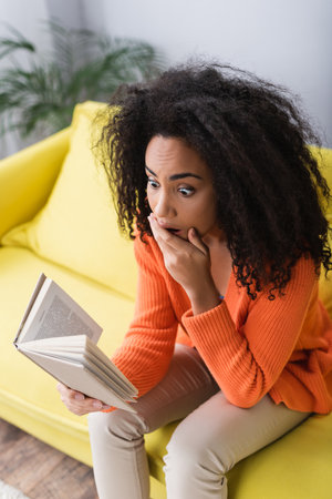 shocked african american woman reading book while sitting on couchの写真素材