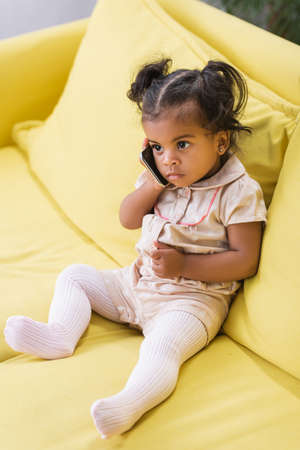toddler african american kid talking on smartphone while sitting on sofaの写真素材