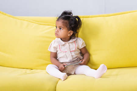 toddler african american child holding smartphone while sitting on sofaの写真素材
